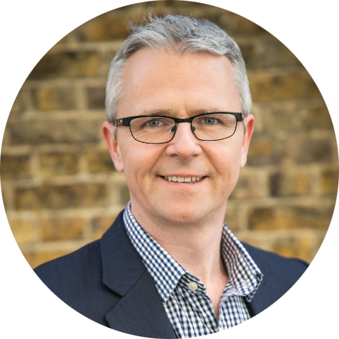 A man with short gray hair and glasses, wearing a dark blazer over a checkered shirt, stands smiling in front of a brick wall, reminiscent of scenes from ITV programs.